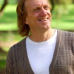Michael White standing under a tree smiling. He is wearing a white t-shirt and a brown cardigan
