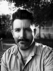 Jake Peterson smiles into the camera, black and white