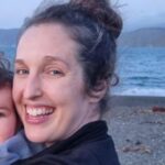 Close up photo of a young white woman with brown hair tied in a high messy bun. She is standing at the beach and smiling. 