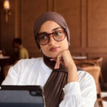 Neha Ayyub sitting in front of a laptop. She wears a white blouse, large eyeglasses and a headscarf or thattam.
