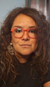 Paola Kolher Salinas. She has long brown hair, is wearing large metal earrings and red rimmed reading glasses