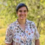 Tanya Newman standing in front of some trees on a sunny day. Tanya is wearing a white shirt with botanical designs