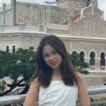 Su Ying. Woman standing in front of an old stone building. Long dark hair and a white top 