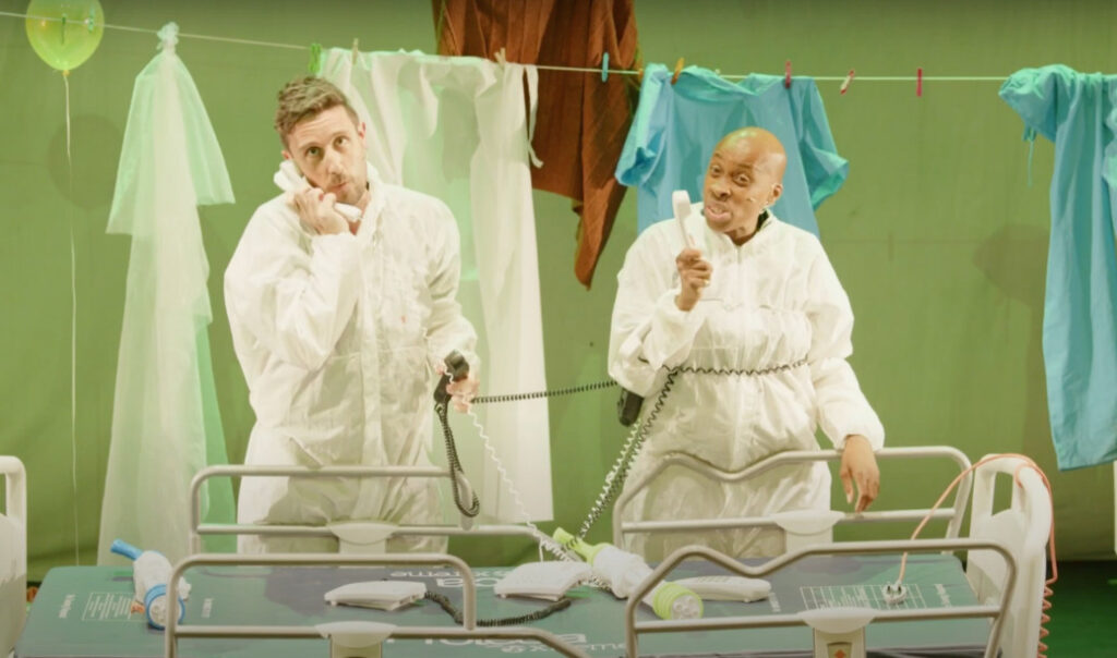 Two actors on stage standing over an empty hospital bed speaking into landline phones