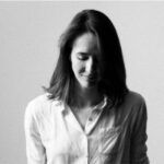 White woman with brown hair and a white un-ironed shirt, looking slightly downward