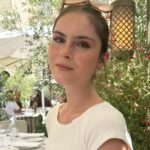 young woman sitting outside at a restaurant, hair tied back and sunglasses on her head. She is wearing a tailored white t-shirt and has a neutral expression