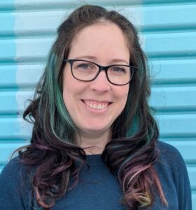 woman with dark hair and glasses, wearing a dark blue top. She has blue, pink and mustard coloured highlights in her hair and is smiling