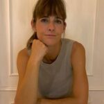 young woman with brown hair and a fringe wearing a grey tank top. Woman is leaning on a table looking thoughtful, with her right fist under her chin