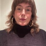 woman with light brown hair and bangs, heavy ring earrings and a brown ribbed turtleneck